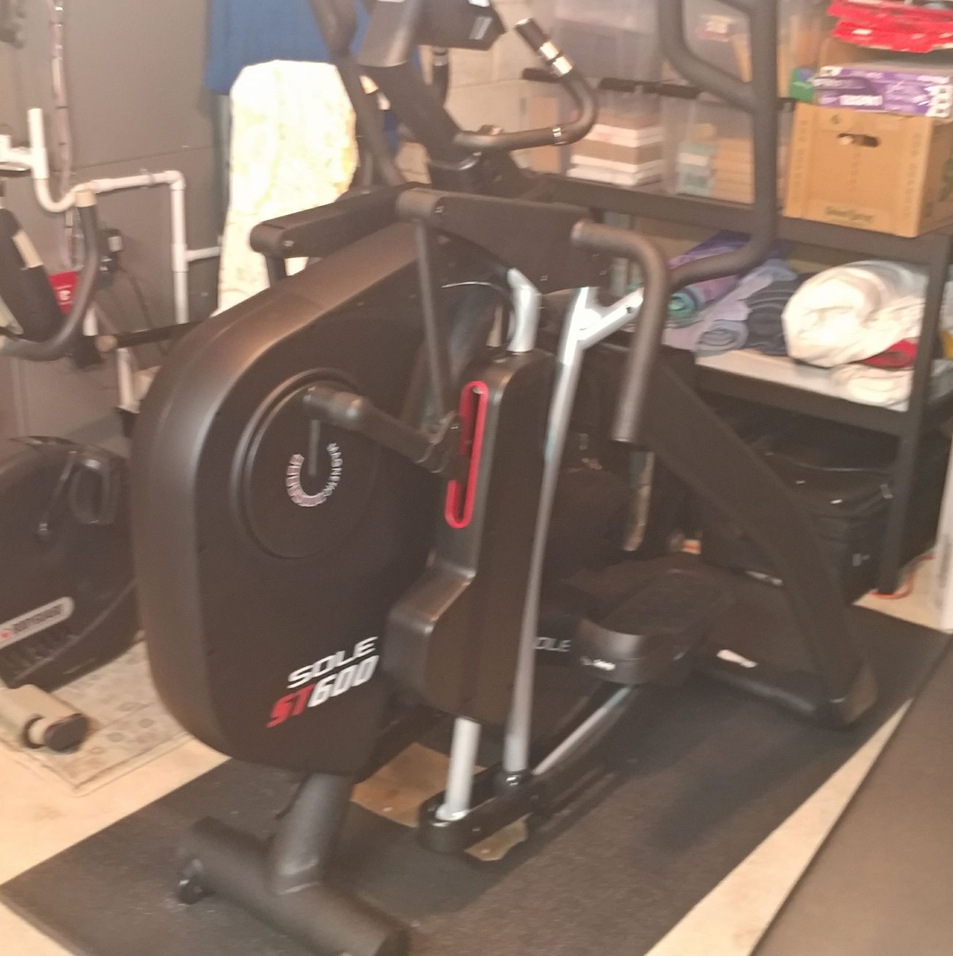 Black exercise bike on a mat in a cluttered storage room or garage.