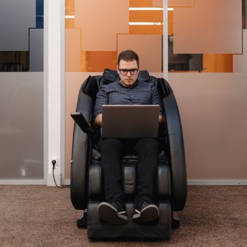 Man using a laptop while seated in a black massage chair in an office lounge