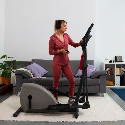 Woman using a black and gray elliptical machine in a living room, wearing a red tracksuit.