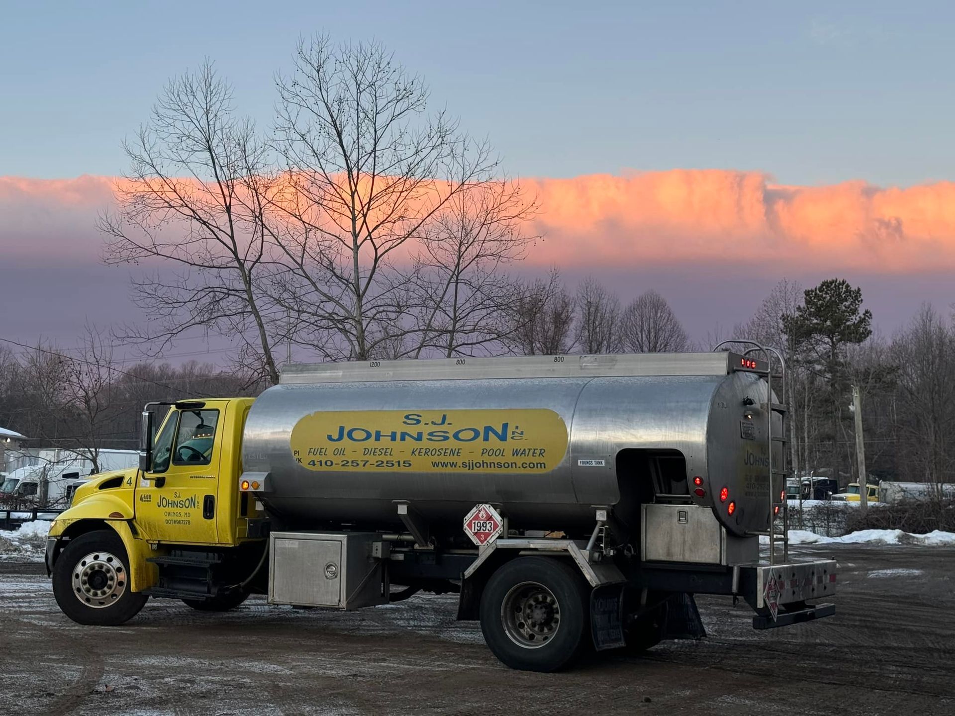 Yellow fuel truck parked outdoors with sunset in the background. Truck has a silver tank and business name.