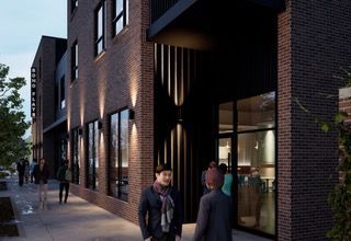 Modern brick building entrance at dusk with people walking on the sidewalk and warm interior lights visible
