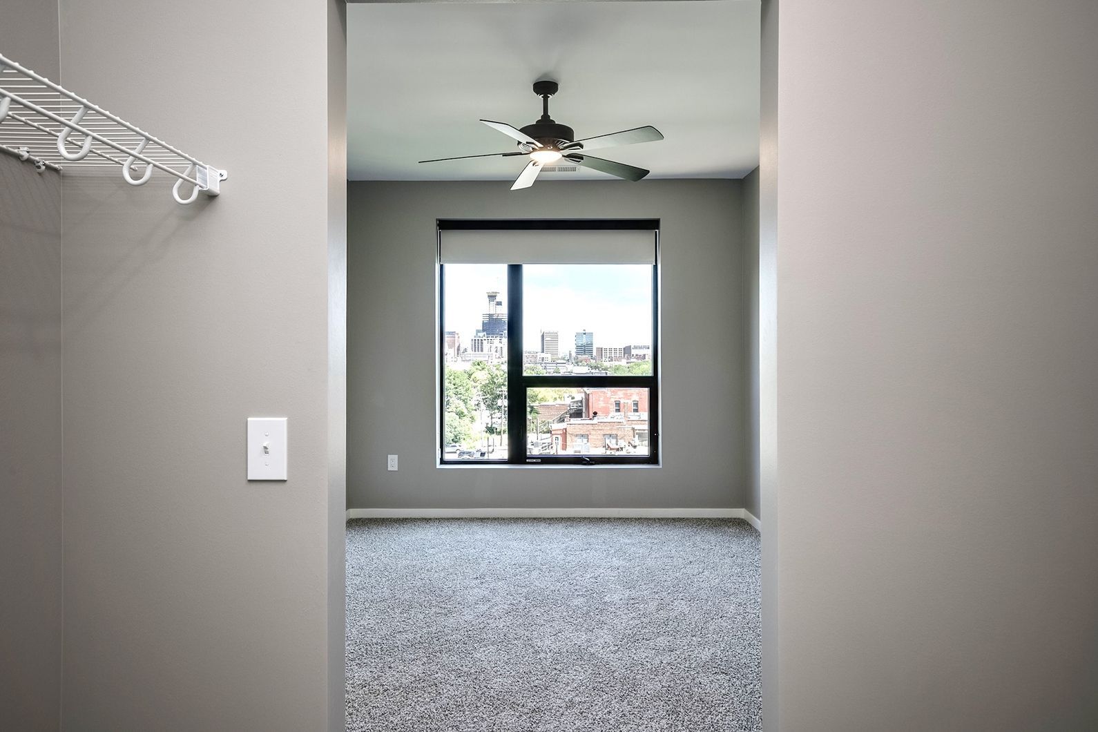 Empty carpeted room with gray walls, ceiling fan, and a window overlooking a city view