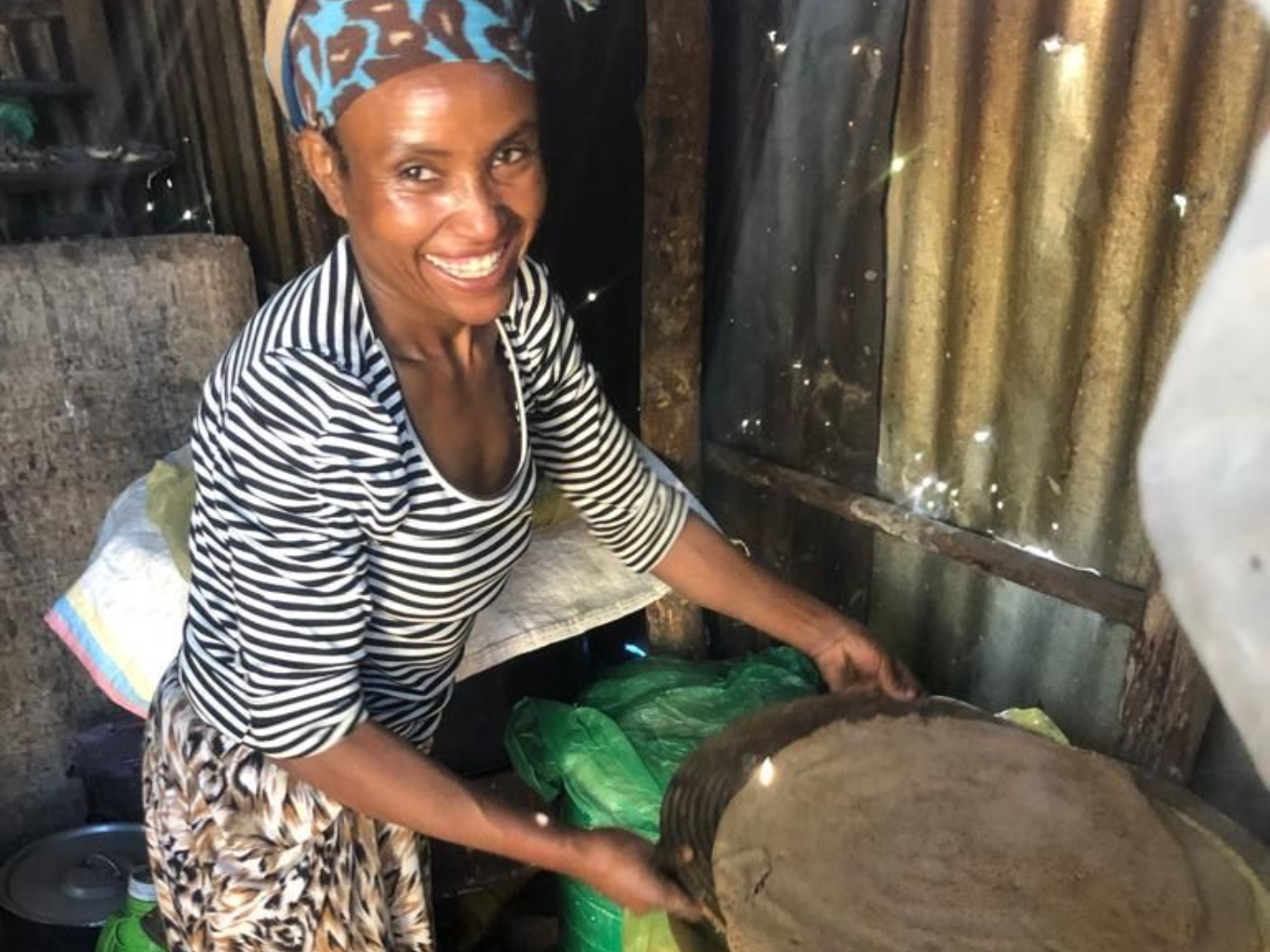 Woman smiling while cooking at a small stove beside a blue drum in a modest kitchen.