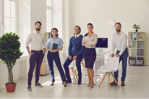 Five businesspeople in an office, some standing, some leaning, having a meeting about salary benchmarking.