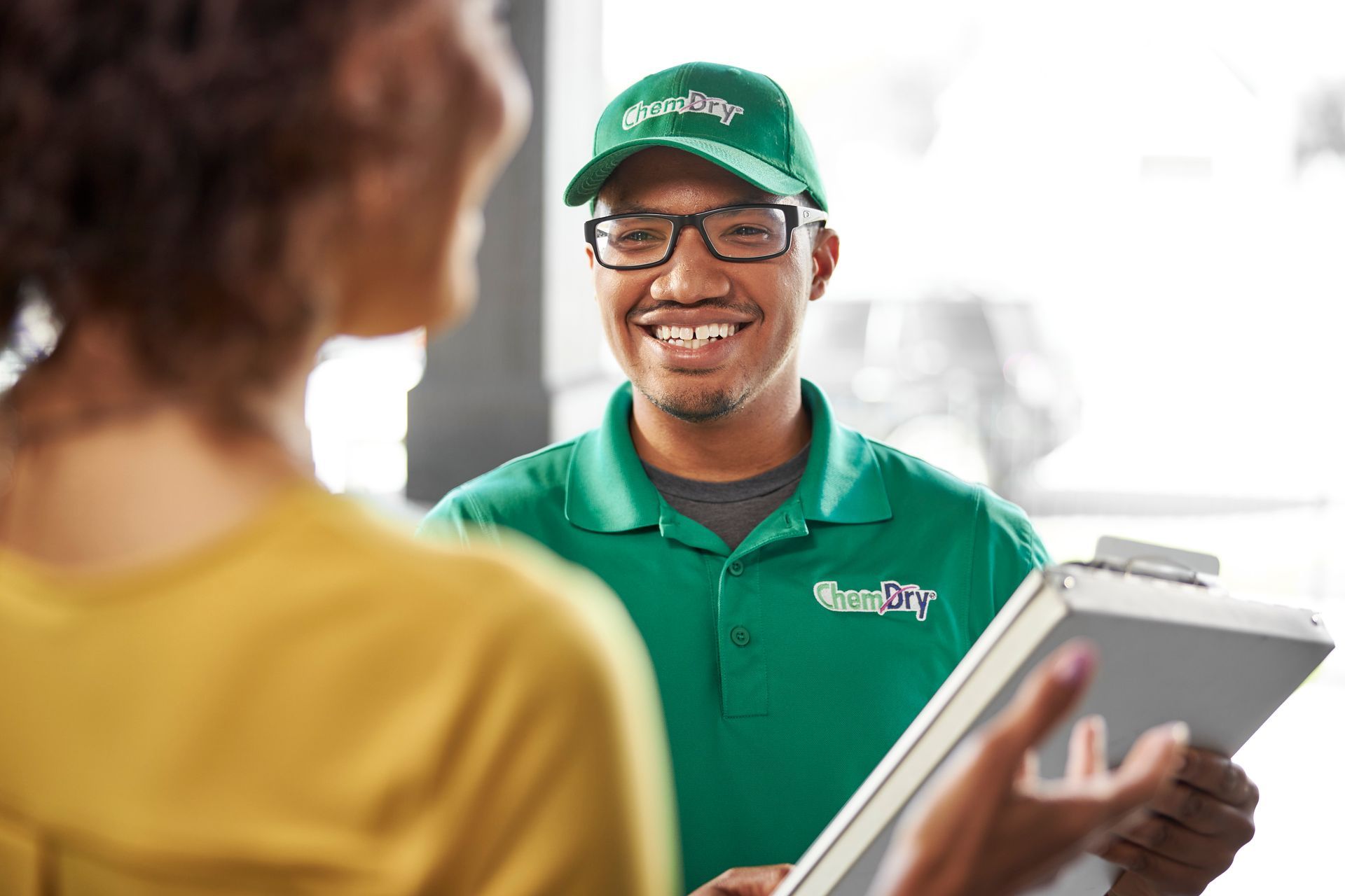 A worker in a green uniform and cap smiles while holding a clipboard and talking to a customer.