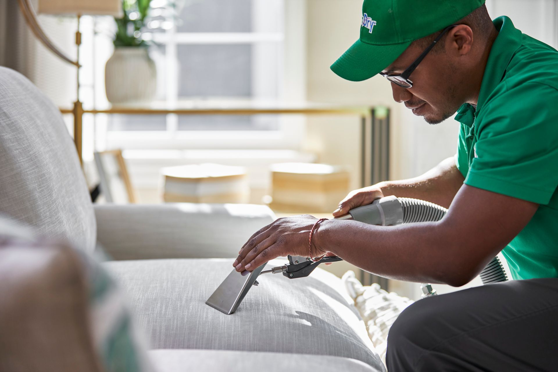 A technician in a green uniform uses a steam cleaner wand on a light-colored fabric sofa.