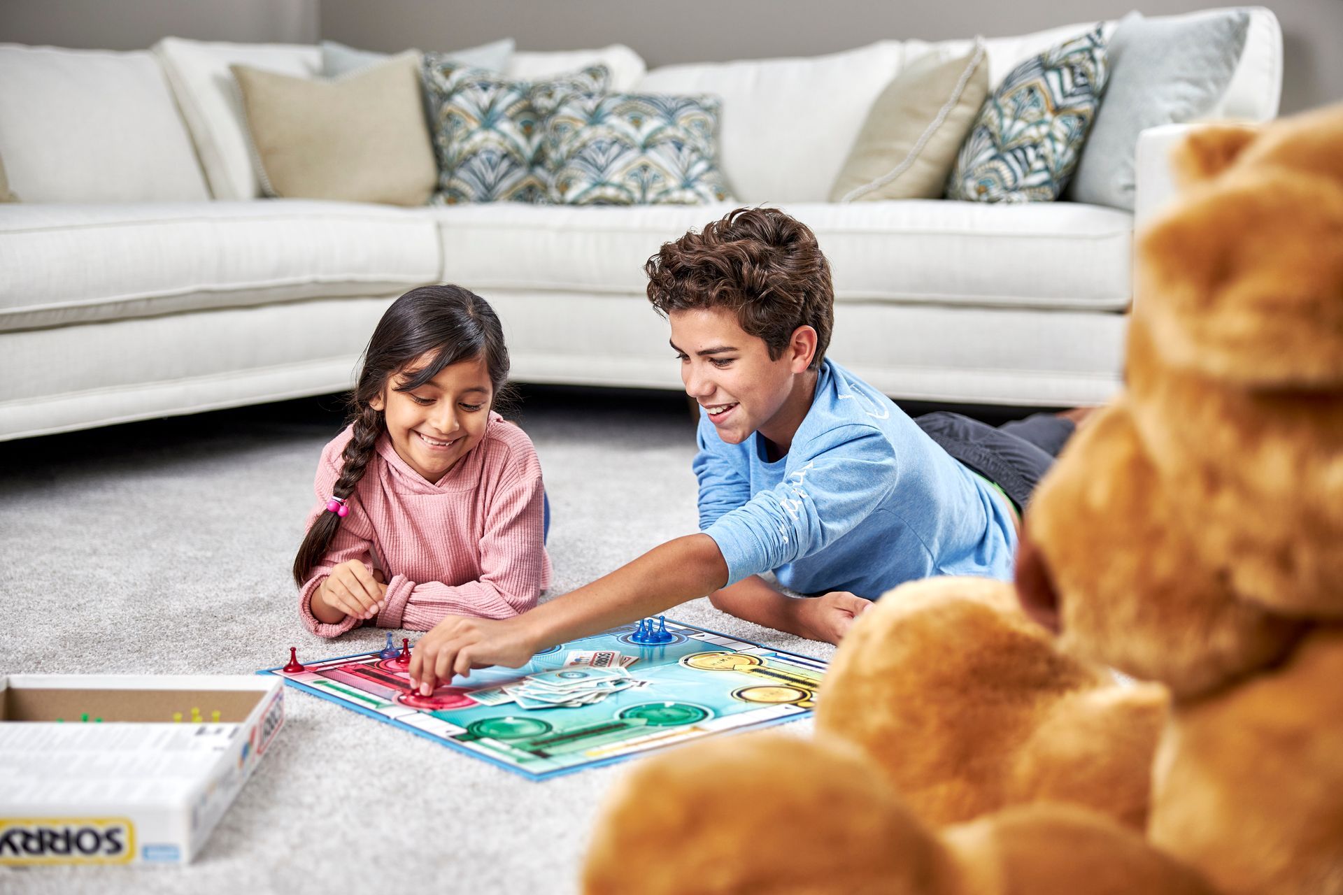 Two children play a board game on a carpeted floor in front of a white sofa, with a large plush teddy bear in the foreground.