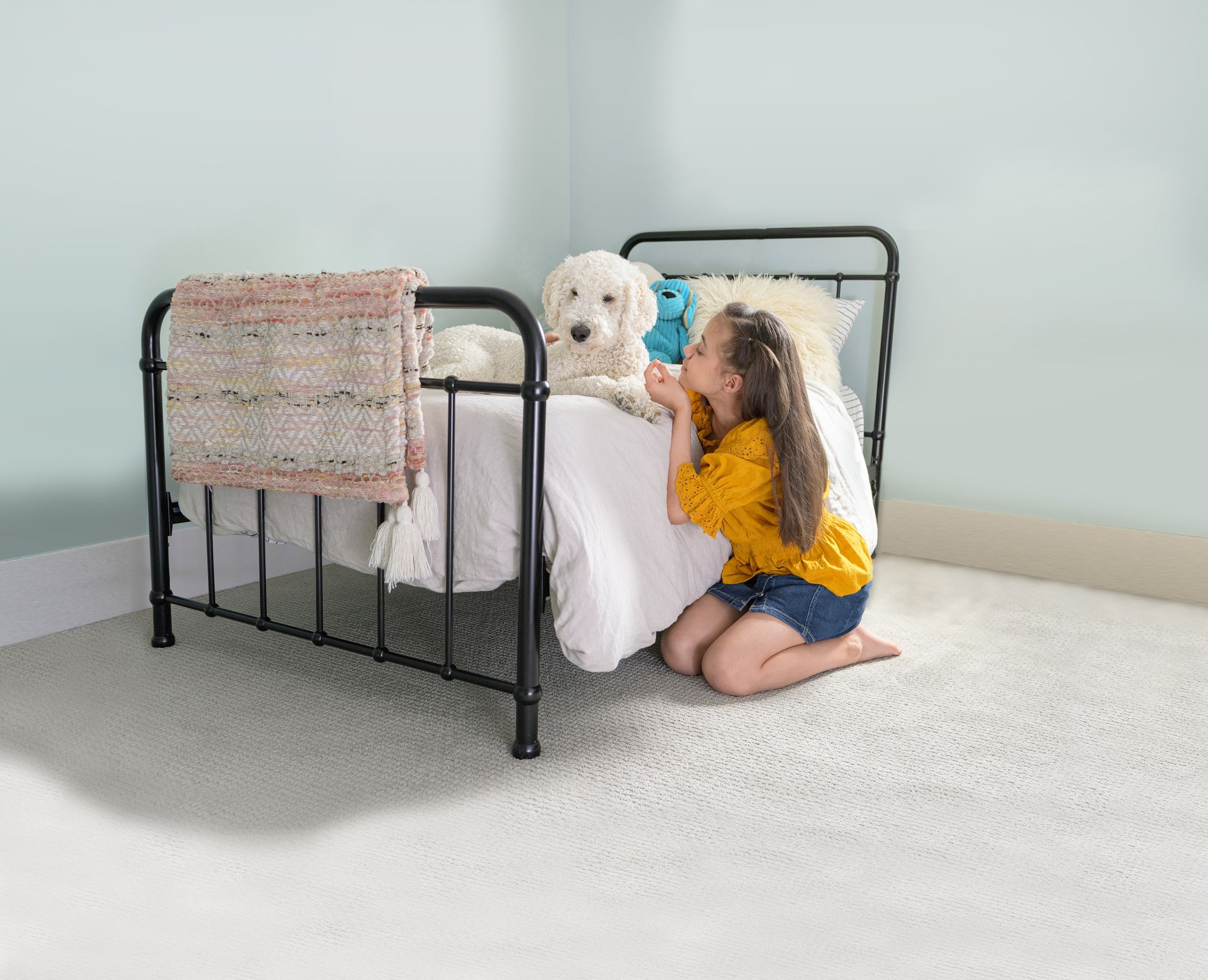 A person in a yellow shirt kneels beside a bed, smiling and petting a fluffy white dog sitting on white bedding.