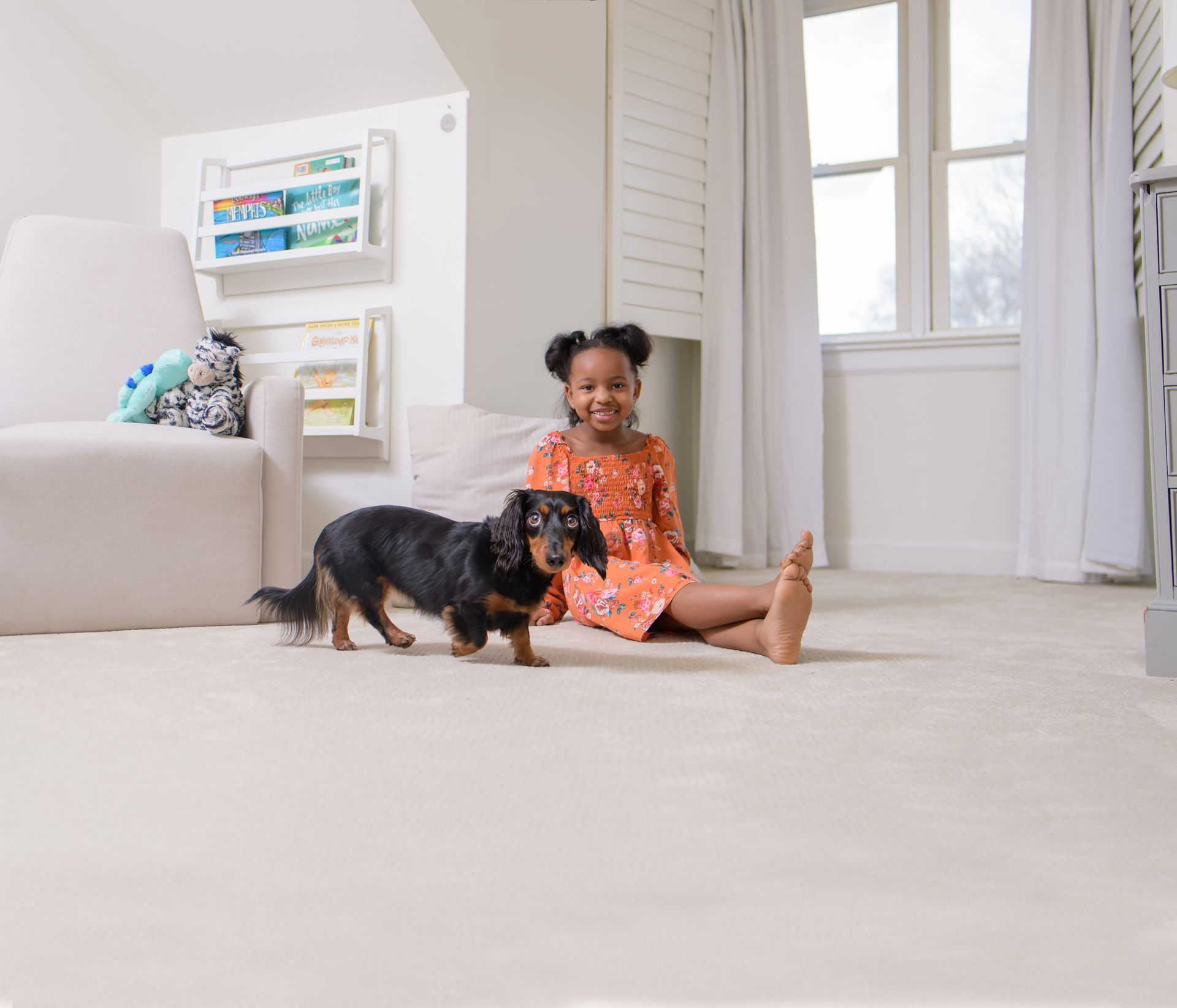 A smiling child sitting on a carpeted floor next to a long-haired dachshund in a bright room with shelves of books.