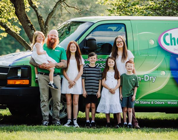 A man, woman, and five children stand in front of a green Chem-Dry van in an outdoor setting.