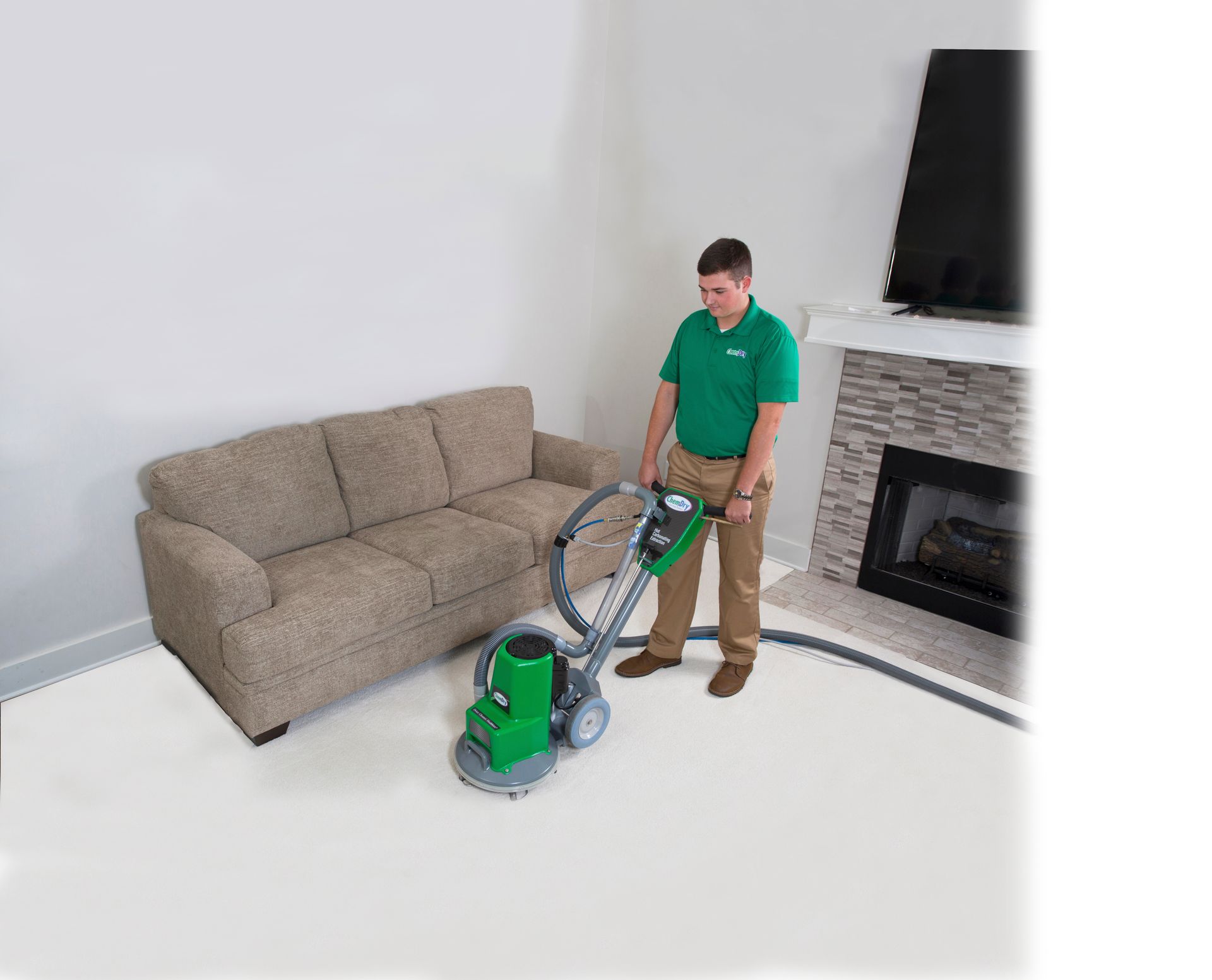 A worker in a green polo shirt cleans a carpeted living room floor with a green and grey industrial floor machine.
