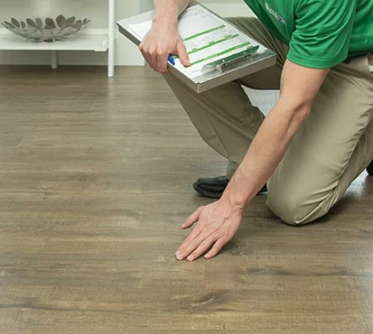 A person in a green shirt and khaki pants kneels on a wood-look floor, holding a clipboard and touching the surface.