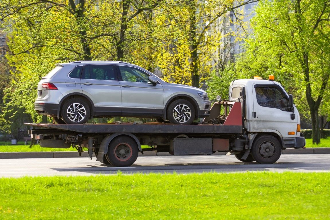 Silver SUV loaded on a flatbed tow truck driving along a road beside green trees.