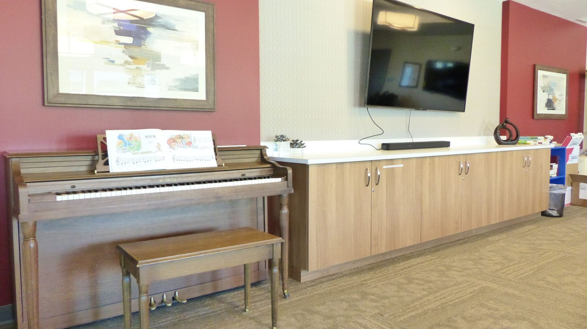 Lobby with a piano, bench, wall-mounted TV, and wood cabinets against a red wall.