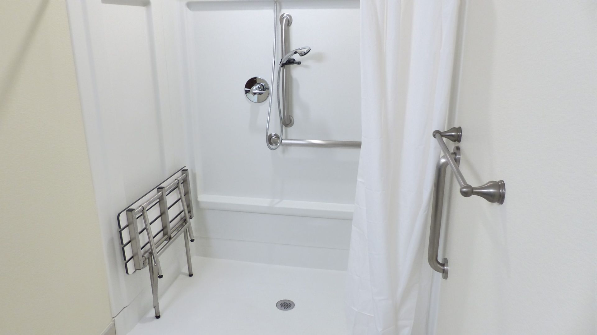 Accessible shower with grab bars, shower chair, and handheld showerhead in a white bathroom.