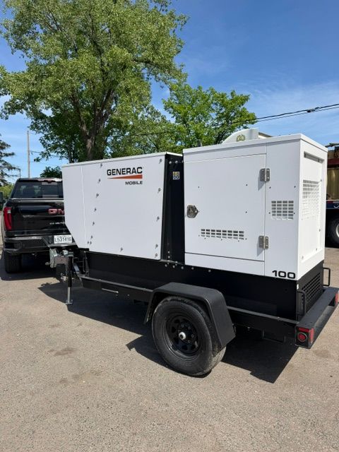 Portable white generator trailer parked on a street beside a black truck under a tree.