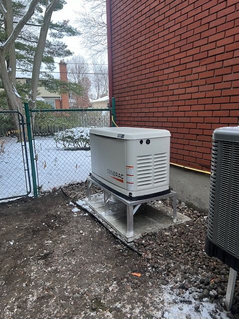 White generator outside a brick building beside a snowy yard and chain-link fence.
