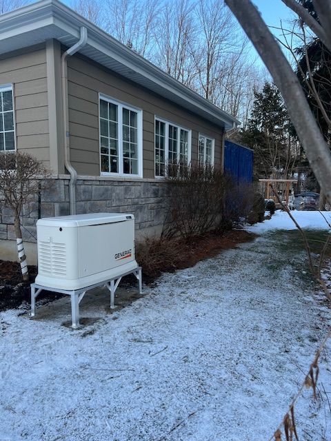 White generator on metal stand beside a beige house in a snowy yard, with bare trees in the background