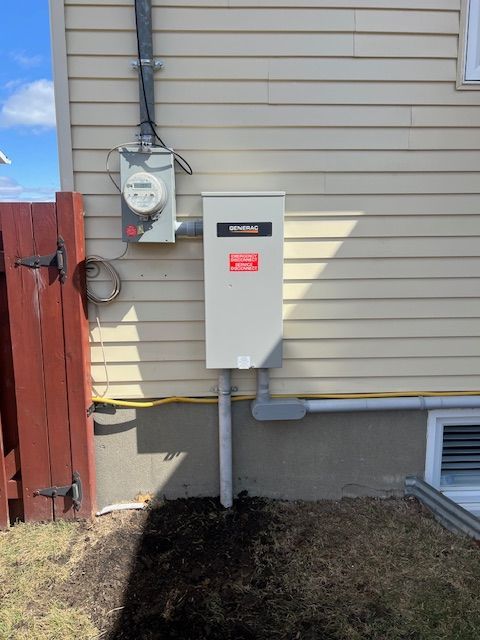 Wall-mounted electrical meter and white utility box on beige house siding beside a red fence.