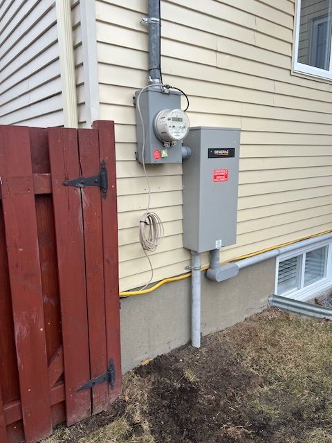 Side of house with electrical meter, gray utility panel, and red wooden gate near the foundation.