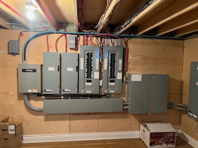 Basement electrical panel with circuit breakers and exposed wiring on a plywood wall.