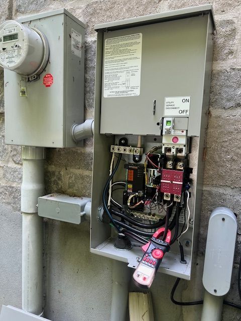 Open electrical meter panel with exposed wiring and a red handheld device attached outdoors on a stone wall