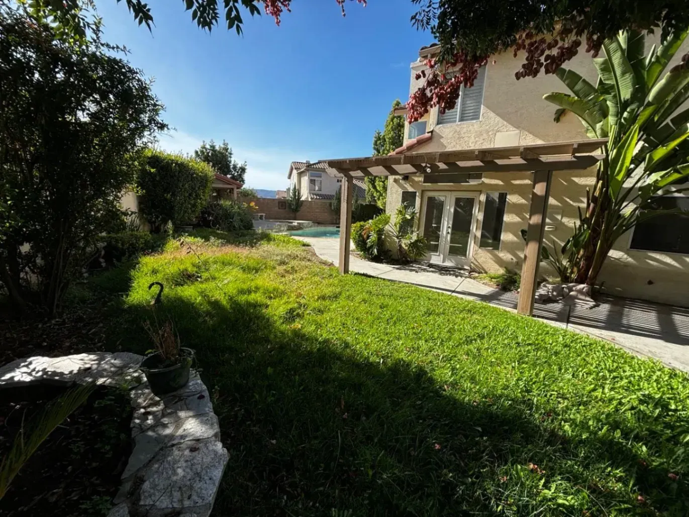Backyard of a house with green lawn, pool, and pergola under a sunny sky.