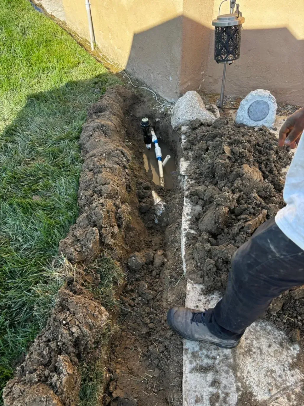 Trench dug near a building. PVC pipe visible. A person's leg and foot are partially in frame, next to the trench.