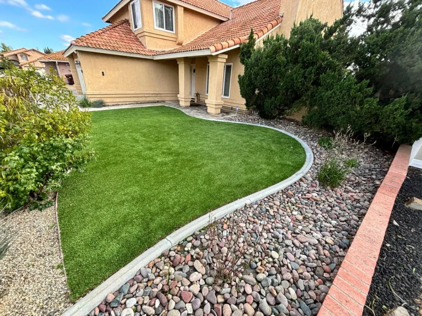 A house with a vibrant green artificial lawn, bordered by decorative rocks and a concrete curb.