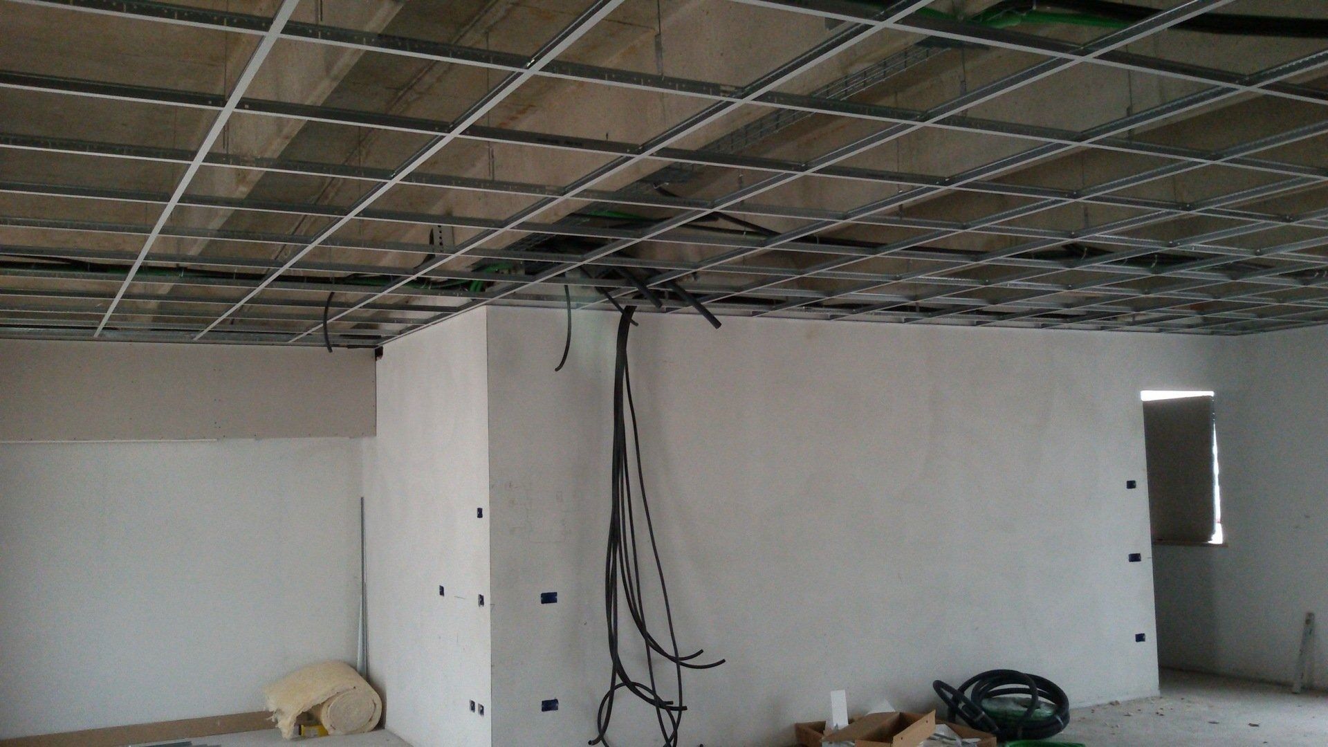 Stanza non rifinita con trave del soffitto a vista, fili penzolanti, pareti bianche e materiali edili sul pavimento.