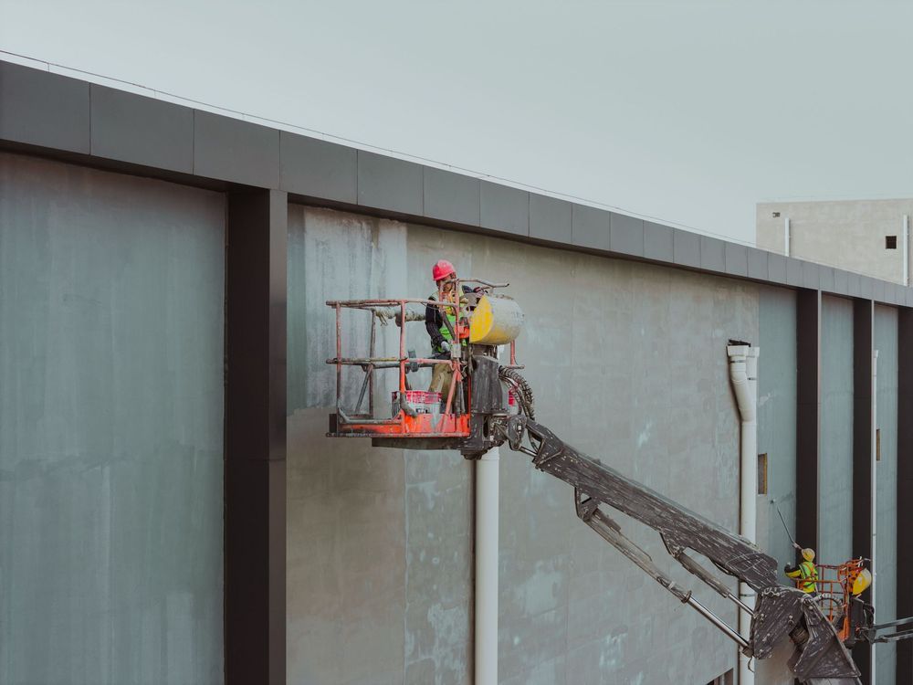 Un operaio in ascensore ripara una parete di un edificio con attrezzi a manico lungo.