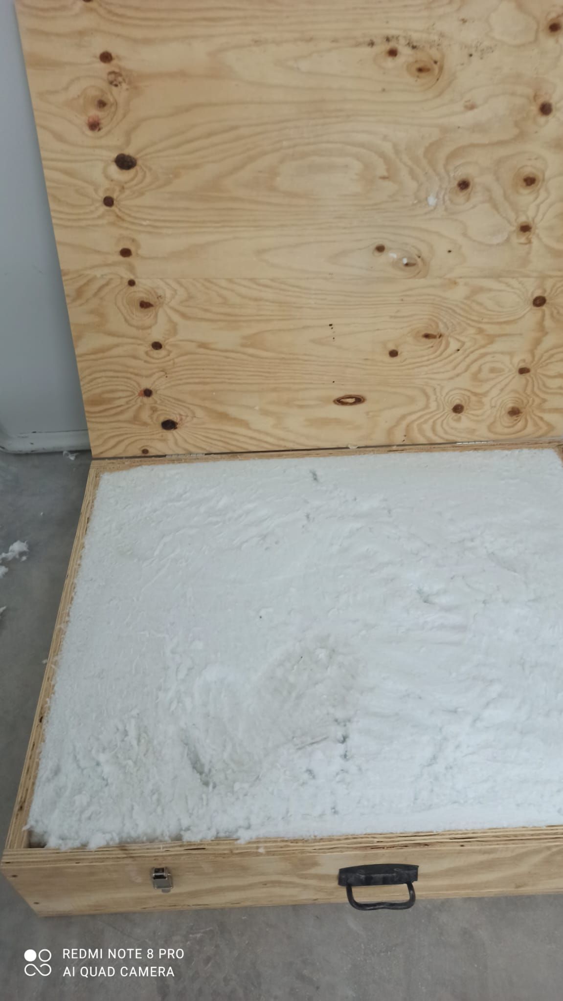Il coperchio aperto di una cassa di legno è posizionato verticalmente dietro una base quadrata riempita con uno spesso strato di schiuma isolante bianca.