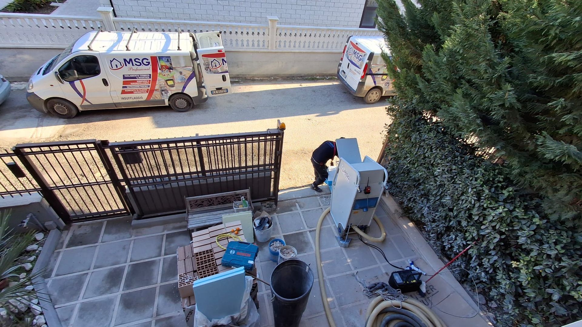 Alcuni operai utilizzano attrezzature industriali vicino all'ingresso di un'abitazione, con due furgoni di servizio bianchi parcheggiati in strada.