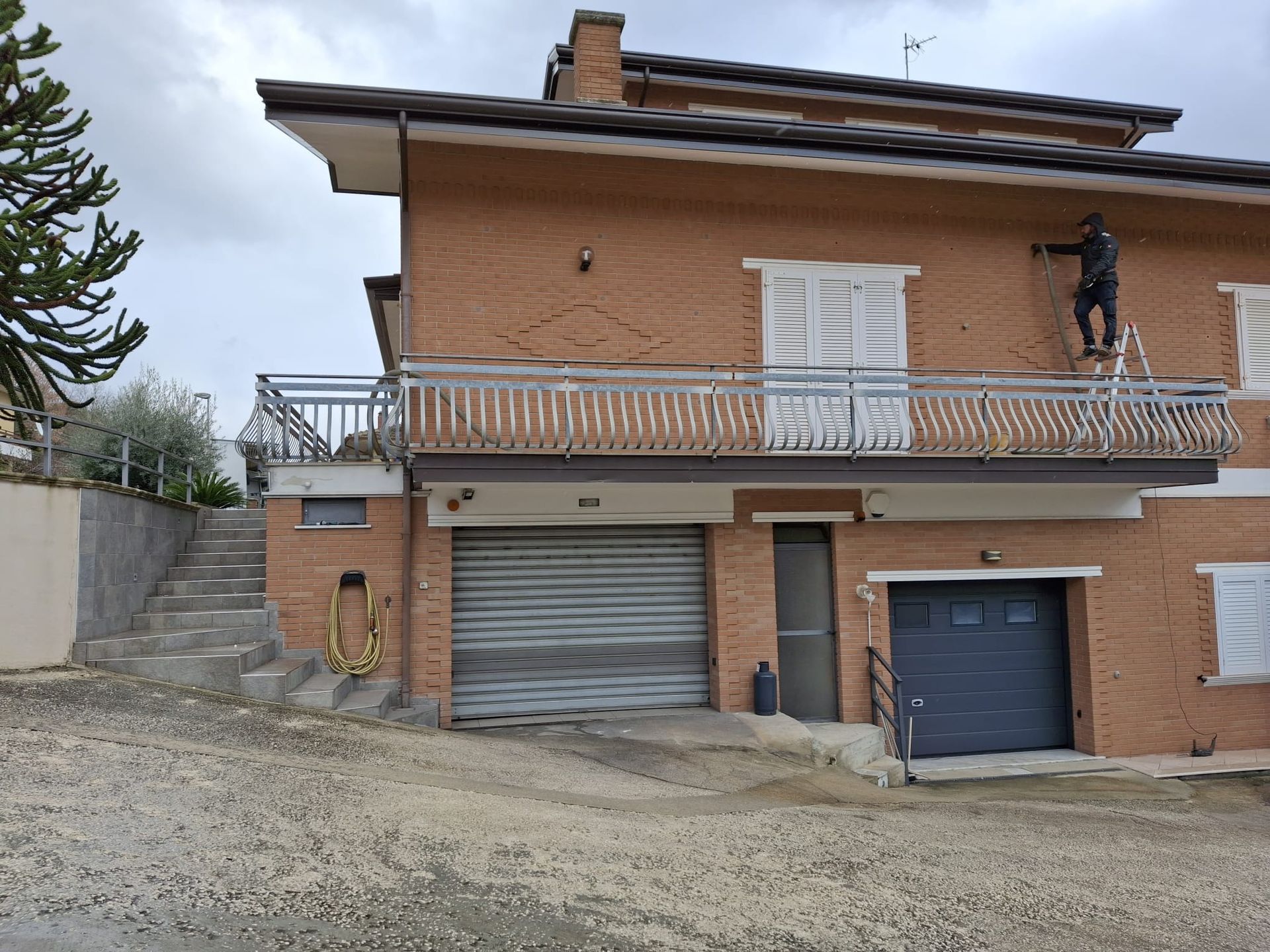 Una persona in piedi su una scala lavora sulla facciata in mattoni di una casa a due piani con balcone e garage al piano terra.