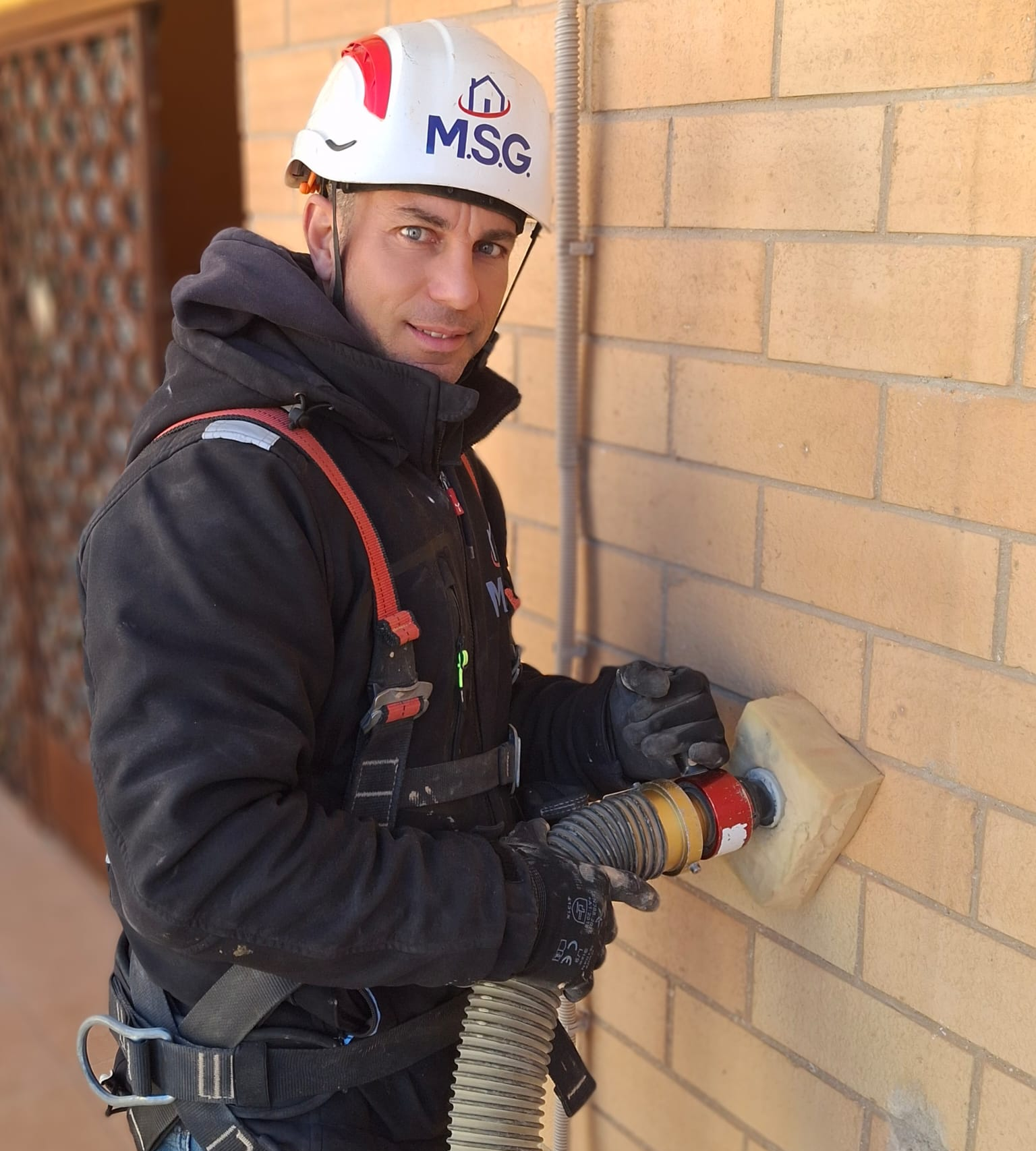 Un operaio, munito di casco e imbracatura di sicurezza, utilizza un tubo aspiratore per pulire un muro di mattoni all'aperto.