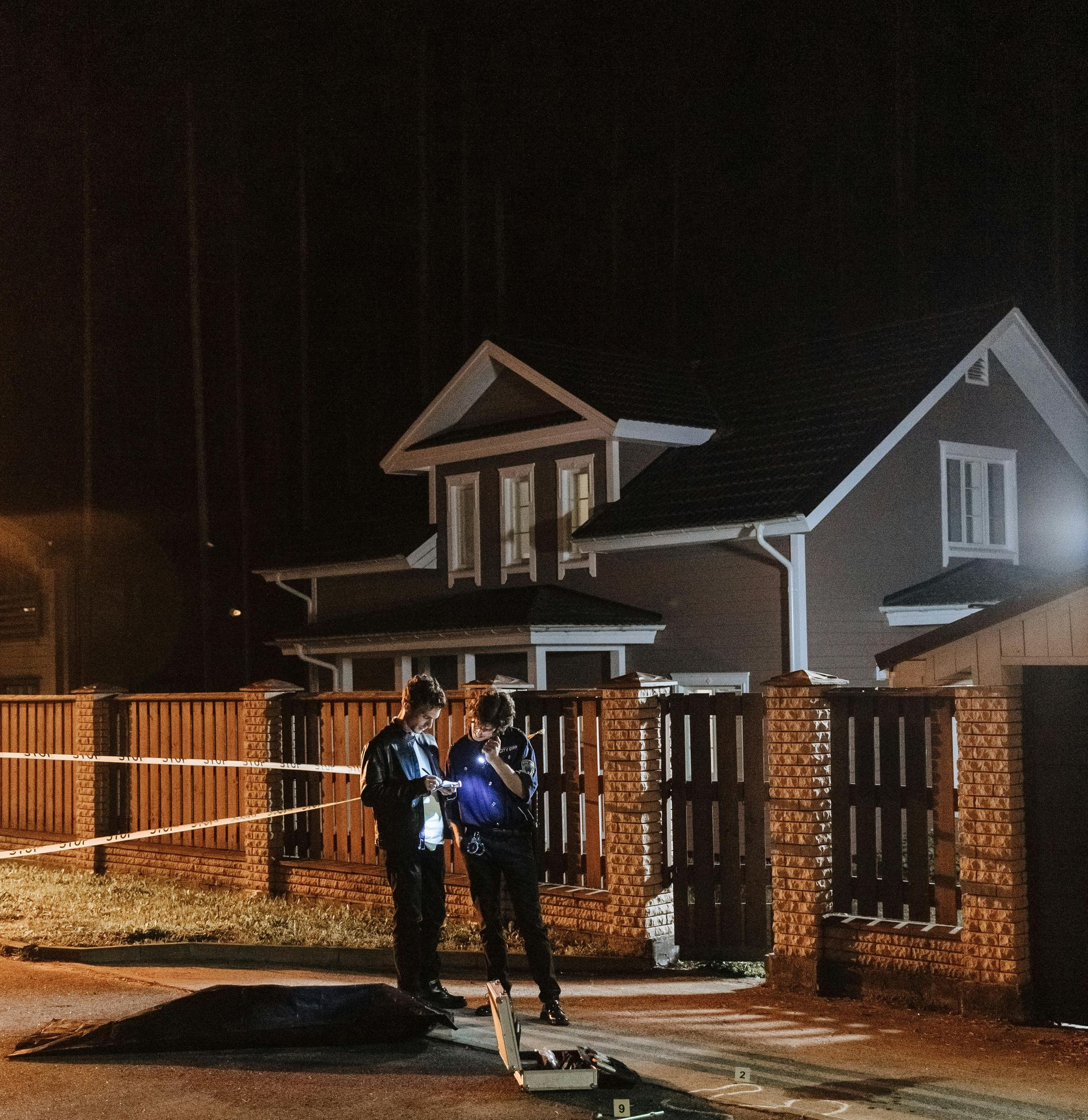 Two people in uniform work at a crime scene at night in front of a house, with police tape and a covered body on the ground.