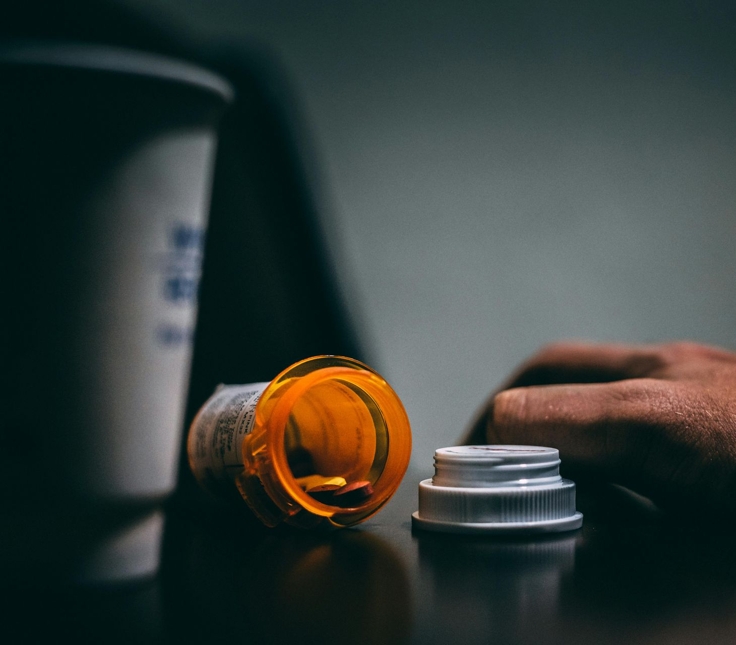 A hand rests near an open orange pill bottle and a cup on a dark surface.