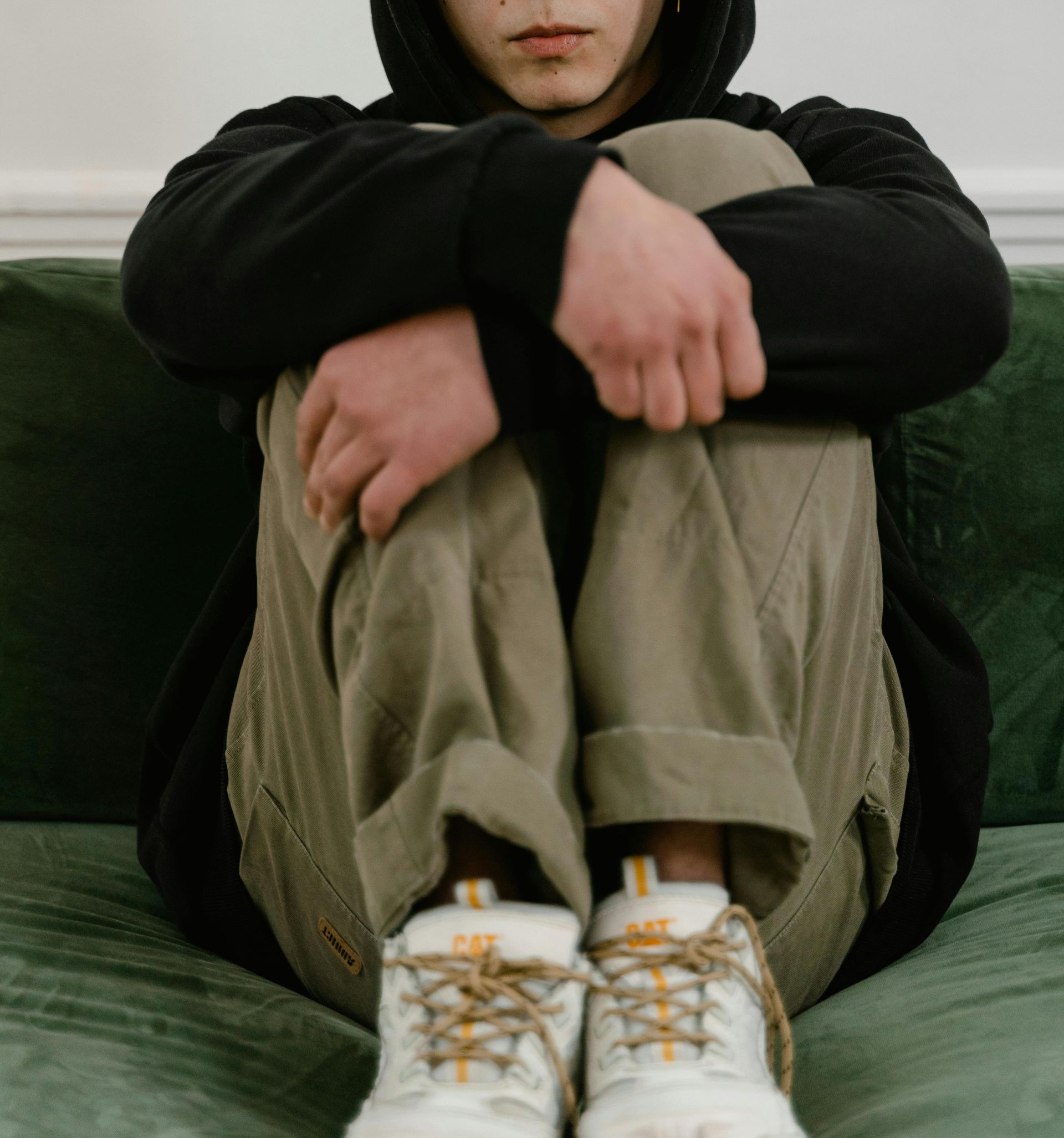 A person in a black hoodie and olive pants sits hunched on a green sofa with their knees tucked toward their chest.
