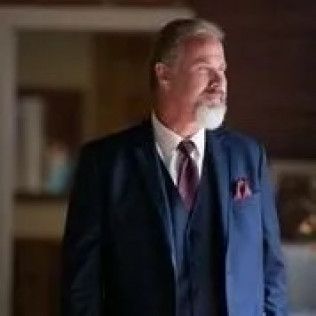 A person with a white beard wearing a blue suit, vest, and tie looks to the side against a dark, blurred background.