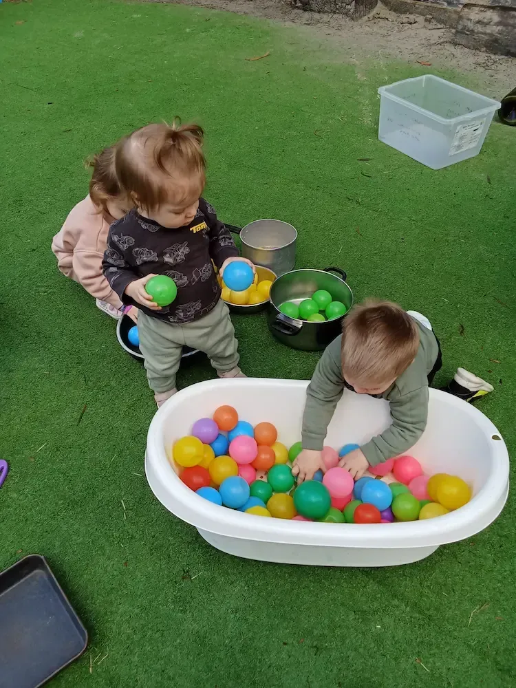Toddlers at a childcare centre engage in sensory play outdoors, exploring a plastic tub filled with colourful plastic balls.