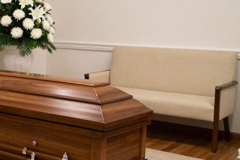 Capilla funeraria con ataúd, arreglos florales blancos y asientos beige.