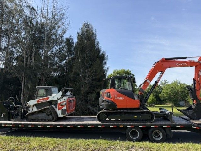 Flatbed trailer carrying a white skid steer and orange mini excavator outdoors under a blue sky