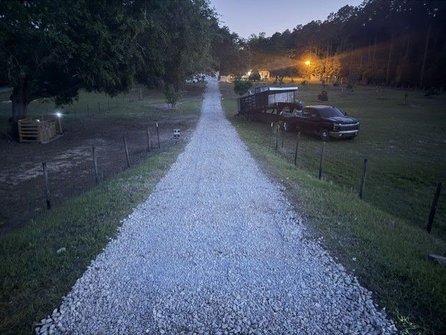 Gravel driveway at dusk between grassy fields, with parked truck and trailer under distant lights