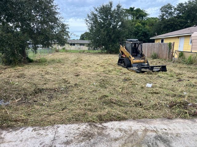 Small yellow bulldozer clearing a grassy yard beside a yellow house under cloudy sky