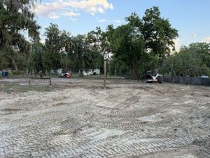 Cleared dirt lot with tire tracks, trees around the edge, and a small white vehicle near the fence