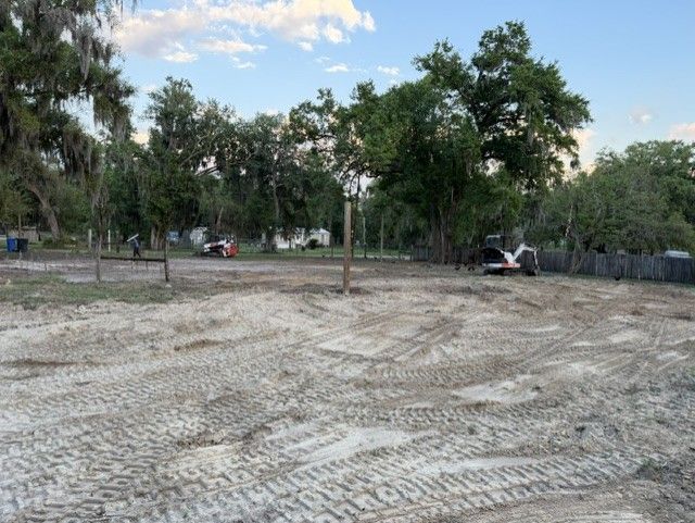 Cleared dirt lot with tire tracks, scattered trees, and parked vehicles under a partly cloudy sky