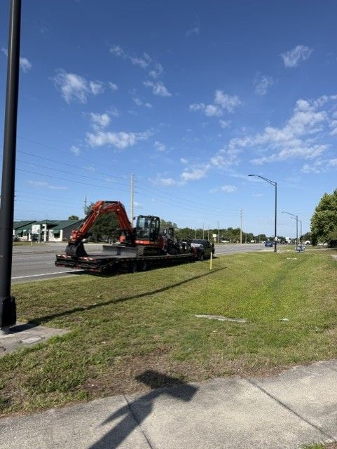 Flatbed truck carrying a large orange excavator on a roadside under a blue sky