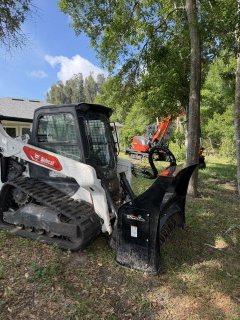 Compact track loader with grapple attachment parked on grass near trees, with an excavator in the background