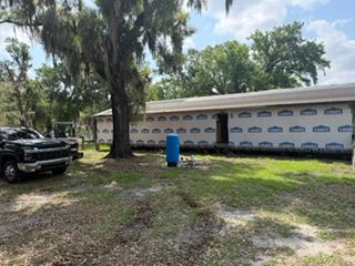 Long white building with blue trim behind trees, parked black truck, and a blue barrel in a grassy yard