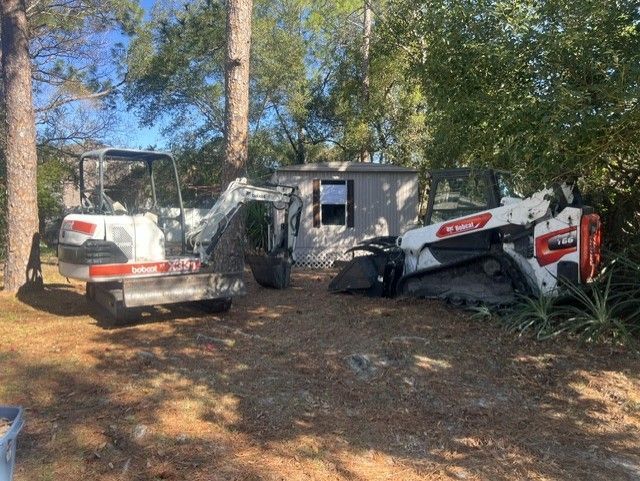 Two construction vehicles parked beside a small house in a wooded yard